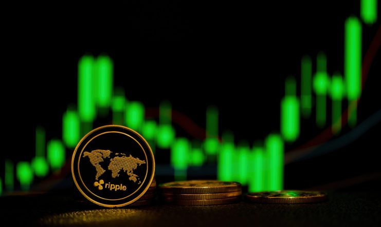 Eine Ripple XRP-Kryptowährungsmünze, die aufrecht vor gestapelten Münzen steht, mit einem grünen Candlestick-Kursdiagramm im unscharfen Hintergrund.