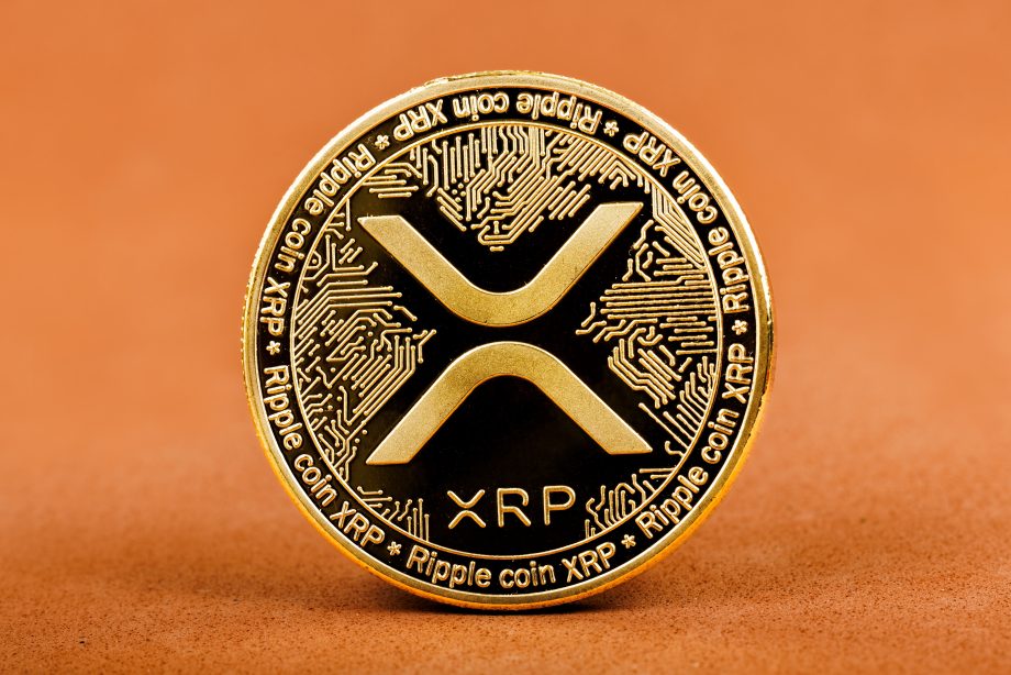 Eine physische Goldmünze mit dem XRP-Logo und dem Schriftzug "Ripple Coin XRP", die aufrecht vor einem soliden orangefarbenen Hintergrund steht - ein passendes Bild, wenn man eine positive XRP-Prognose betrachtet.