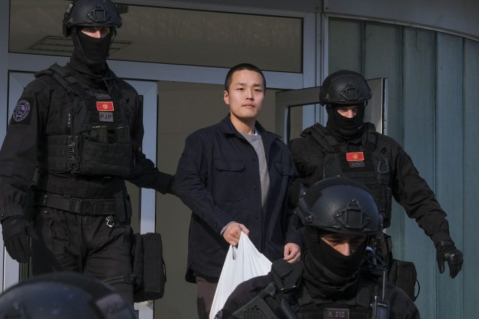 Ein asiatischstämmiger Mann verlässt in Begleitung vermummter Polizisten ein Gebäude in Montenegro.