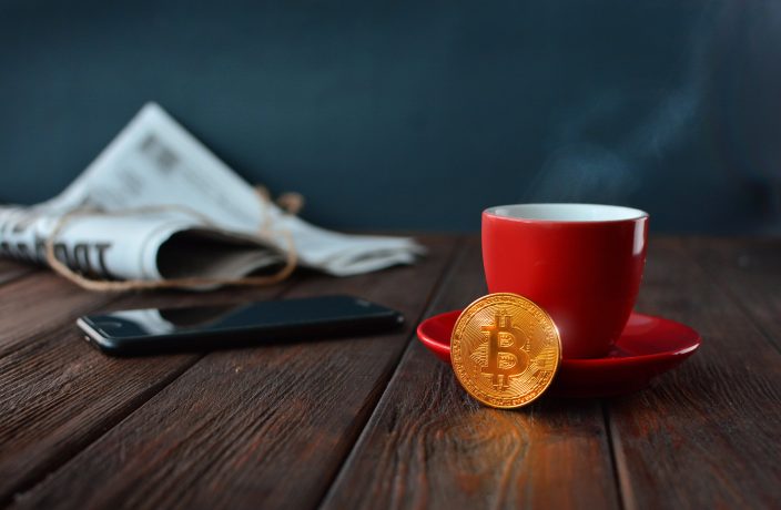 Eine rote Kaffeetasse mit Untertasse steht auf einem Holztisch mit einer goldenen Bitcoin-Münze, die die Welt der Kryptowährung symbolisiert, neben einem Smartphone und einer zusammengerollten Zeitung im Hintergrund.