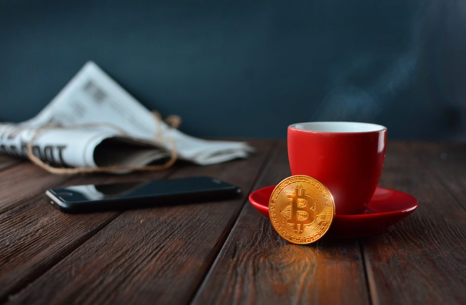 Auf einem Holztisch ist eine Bitcoin-Münze vor einer roten Tasse zu sehen. Dahinter liegt ein Smartphone und eine Zeitung.