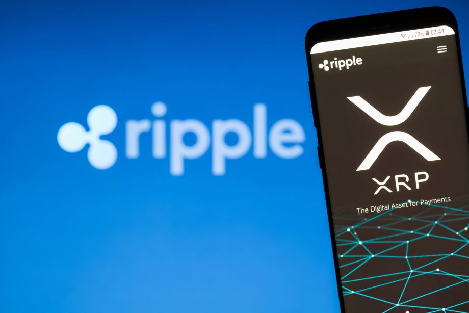 Auf einem Smartphone wird das Ripple-XRP-Logo mit dem Text „The Digital Asset for Payments“ vor einem leicht verschwommenen Ripple-Hintergrund angezeigt.