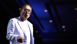 Robert Kiyosaki spricht auf einer Bühne im hellen Anzug vor blauem Hintergrund
