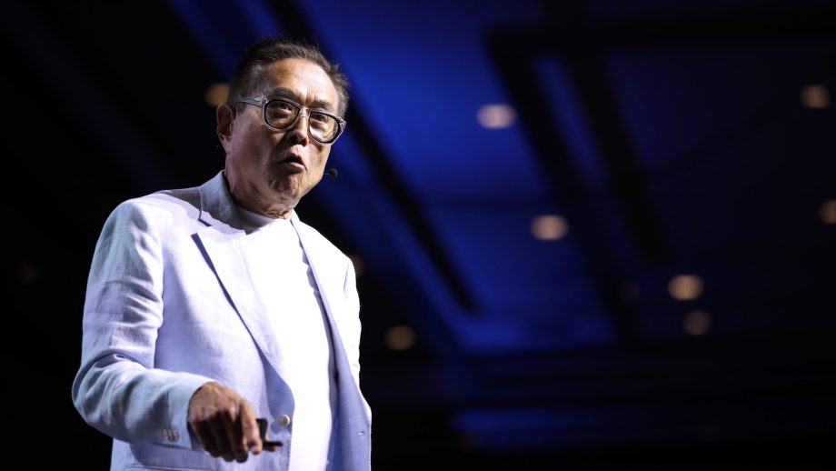 Robert Kiyosaki spricht auf einer Bühne im hellen Anzug vor blauem Hintergrund