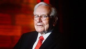 Warren Buffett, ein älterer Mann in Anzug und roter Krawatte, sitzt vor einem roten Hintergrund, blickt leicht zur Seite und lächelt.
