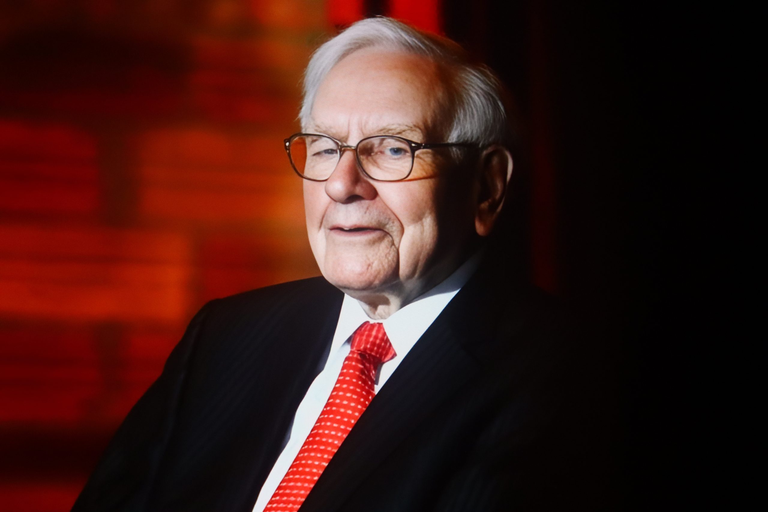 Warren Buffett, ein älterer Mann in Anzug und roter Krawatte, sitzt vor einem roten Hintergrund, blickt leicht zur Seite und lächelt.