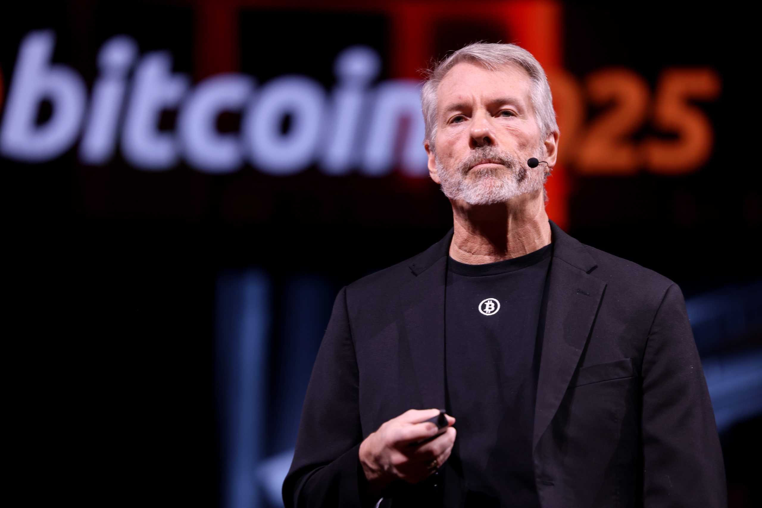 Ein grauhaariger, bärtiger Mann steht auf einer Bühne. Hinter ihm steht geschrieben "Bitcoin 2025".