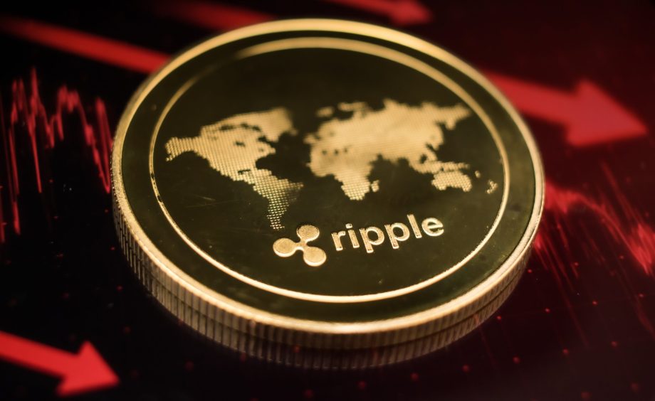 Eine goldene XRP-Münze mit dem Ripple-Logo und einer Weltkarte ruht auf einer dunklen Oberfläche, die von roten Finanzdiagrammlinien beleuchtet wird.