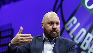 Ein glatzköpfiger Mann im Anzug gestikuliert mit seiner rechten Hand, während er über Bitcoin spricht. Er sitzt vor einem violetten Hintergrund, auf dem ein Teil des Textes zu sehen ist.