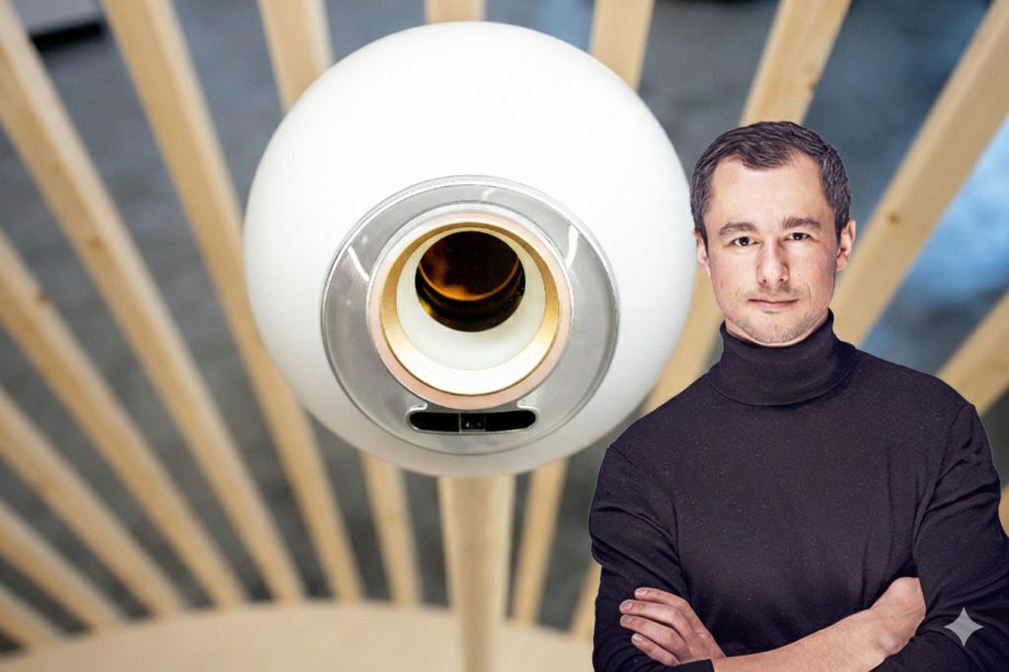 Auf dem Bild ist ein Iris Scanner von Worldcoin abgebildet. Im Hintergrund sieht man mehrere Holzsträben. Rechts steht ein Mann im schwarzen Rollkragenpullover.