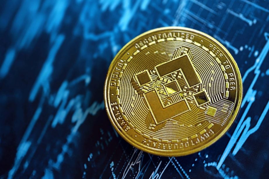 Eine goldene BNB Binance-Münze ruht auf einem blauen Hintergrund, auf dem Finanzcharts und Datengrafiken zu sehen sind.