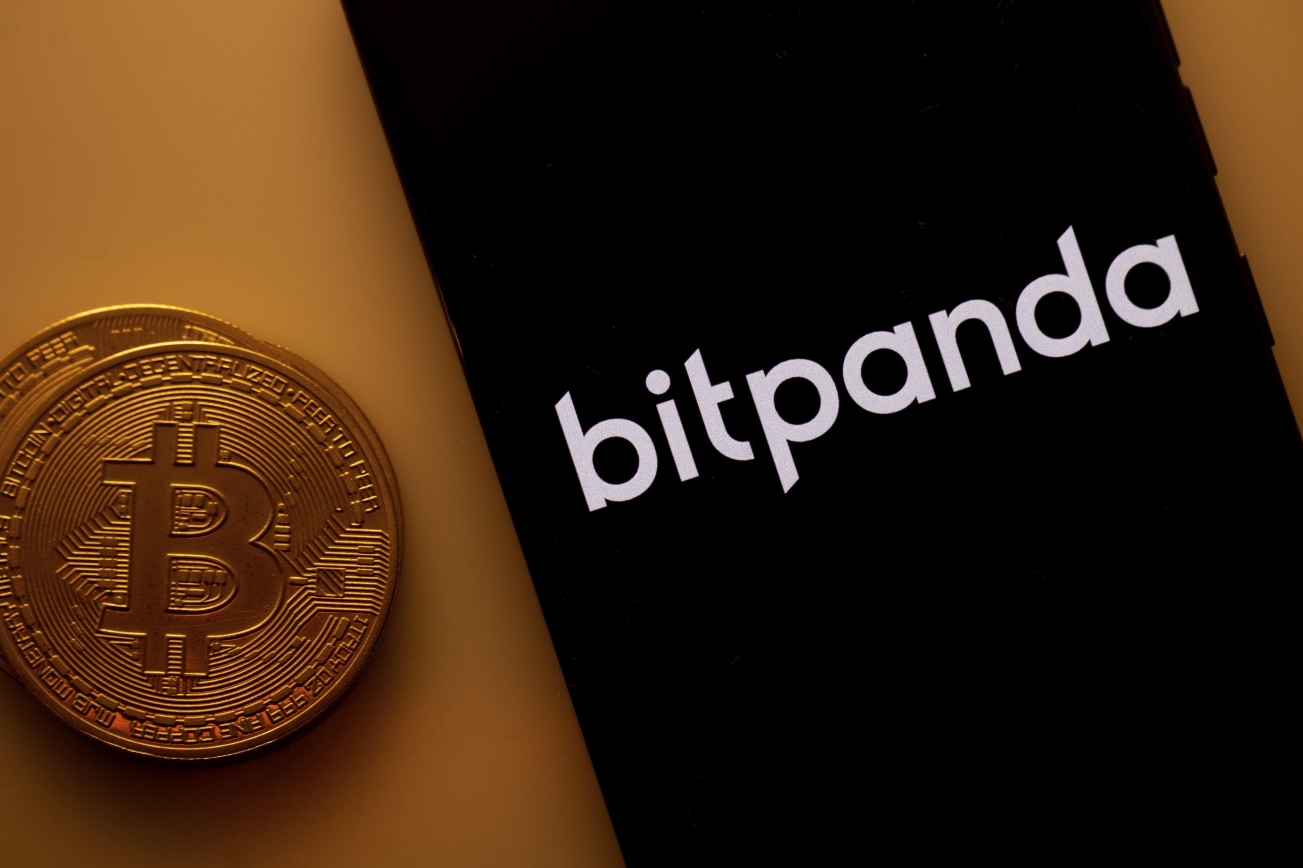 Bitpanda Logo auf dem Bildschirm eines Smartphones neben einer Bitcoin Münze.