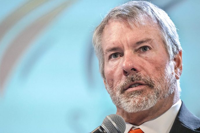 Ein Mann mittleren Alters mit grauen Haaren und Bart spricht in ein Mikrofon über Bitcoin. Er trägt einen Anzug und eine orangefarbene Krawatte, der Hintergrund ist hellblau und verschwommen.