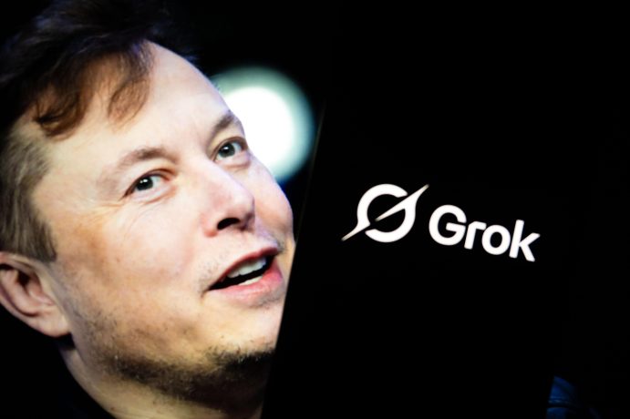 Elon Musk steht neben einem Bildschirm, auf dem das Grok-Logo mit weißem Text und einem stilisierten Symbol auf schwarzem Hintergrund zu sehen ist, daneben ein dezentes Bitcoin-Symbol.