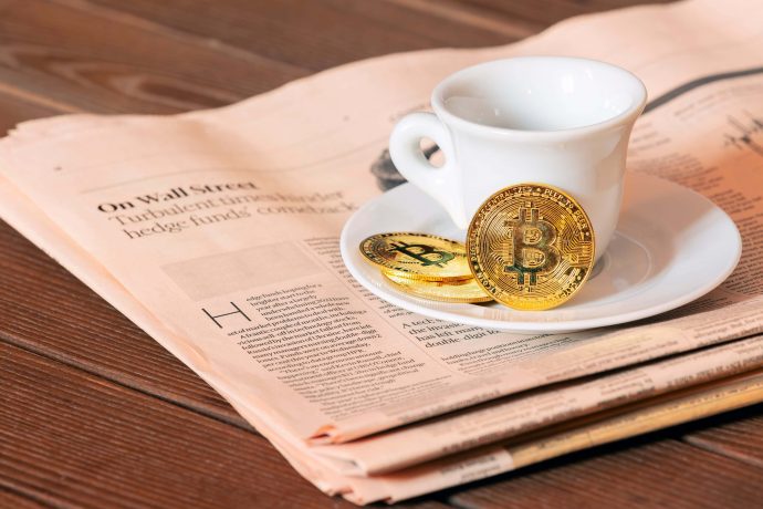 Eine weiße Kaffeetasse steht auf einer Untertasse auf einer Zeitung, daneben liegen zwei physische Bitcoin-Münzen und ein Hauch von Krypto-Intrige.