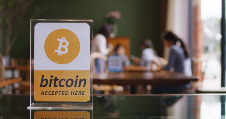 Auf einem Schild steht "bitcoin accepted here", dahinter sind Menschen zu sehen
