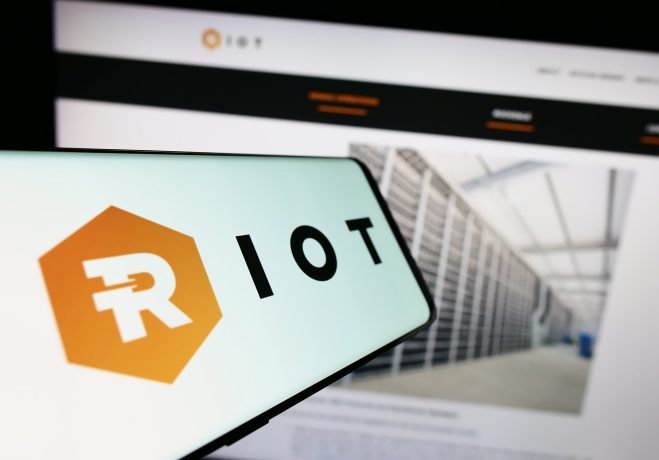 Auf einem Smartphone wird das Logo von Bitcoin Miner Riot Platforms vor einem Computerbildschirm angezeigt, auf dem die Website des Unternehmens mit einem Bild eines Serverraums dargestellt ist.