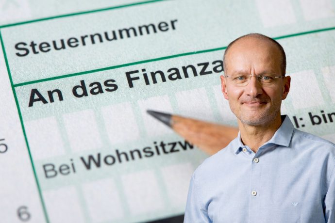Montage mit einem Steuerformular mit der Aufschrift „An das Finanzamt“ im Hintergrund und einem Mann im hellblauen Hemd im rechten unteren Bildbereich. Das Bild steht symbolisch für das Thema Steuer, Steuererklärung und steuerliche Pflichten.