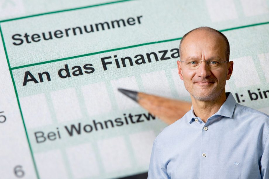 Montage mit einem Steuerformular mit der Aufschrift „An das Finanzamt“ im Hintergrund und einem Mann im hellblauen Hemd im rechten unteren Bildbereich. Das Bild steht symbolisch für das Thema Steuer, Steuererklärung und steuerliche Pflichten.