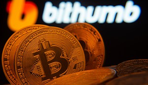 Drei rot gefärbte Bitcoin-Münzen stehen vor einem verschwommen erkennbaren Bithumb-Logo als Hintergrund