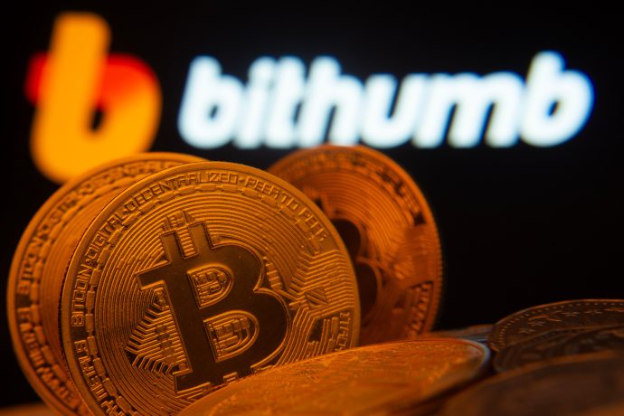 Drei rot gefärbte Bitcoin-Münzen stehen vor einem verschwommen erkennbaren Bithumb-Logo als Hintergrund