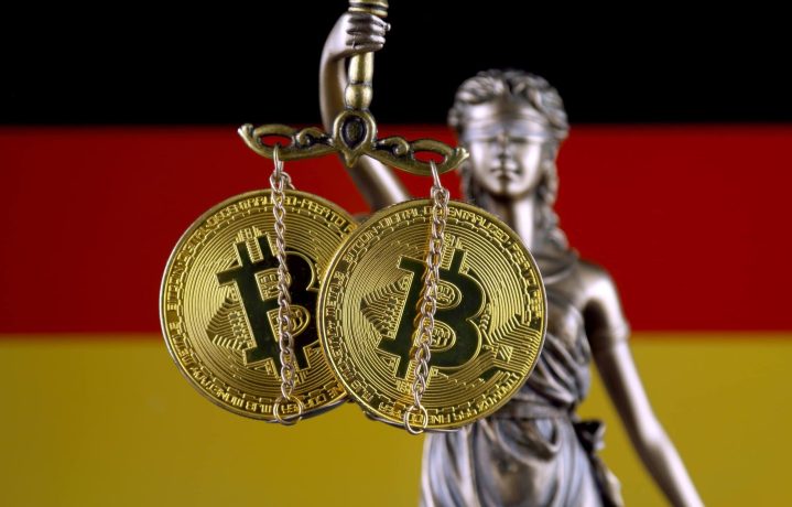 Wem wird der Bitcoin-Schatz zugesprochen? Zwei Bitcoin-Münzen werden von einer Justizia-Figur gehalten, während man im Hintergrund die deutsche Flagge sieht.