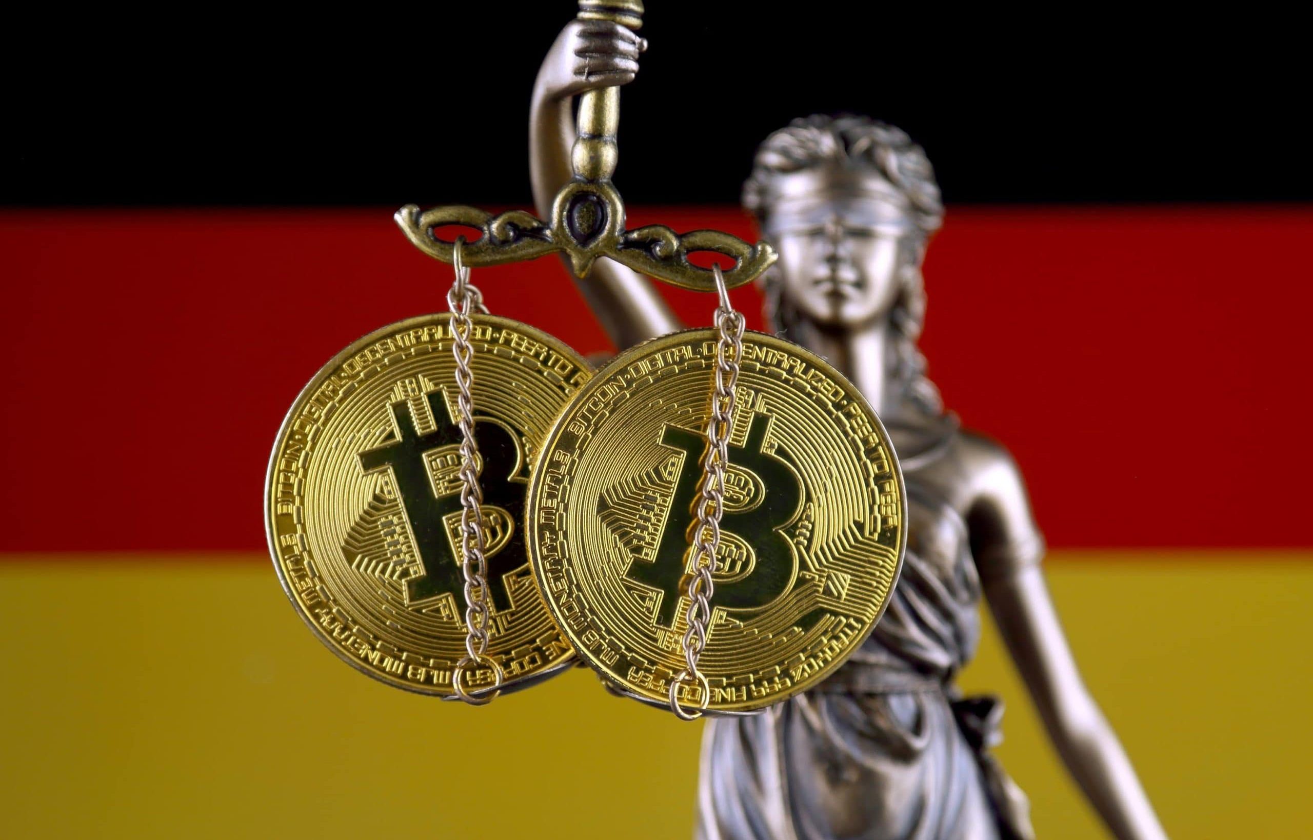 Zwei Bitcoin-Münzen werden von einer Justizia-Figur gehalten, während man im Hintergrund die deutsche Flagge sieht.