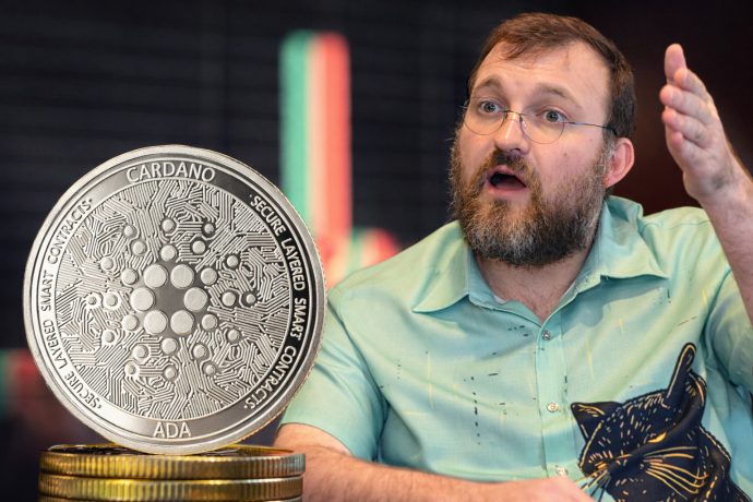 Das Bild zeigt eine Cardano-Münze. Neben ihr hebt Gründer Charles Hoskinson seinen linken Arm.