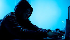 Eine dunkel gekleidete Person sitzt im Schatten vor einem Laptop vor einem blauen Hintergrund