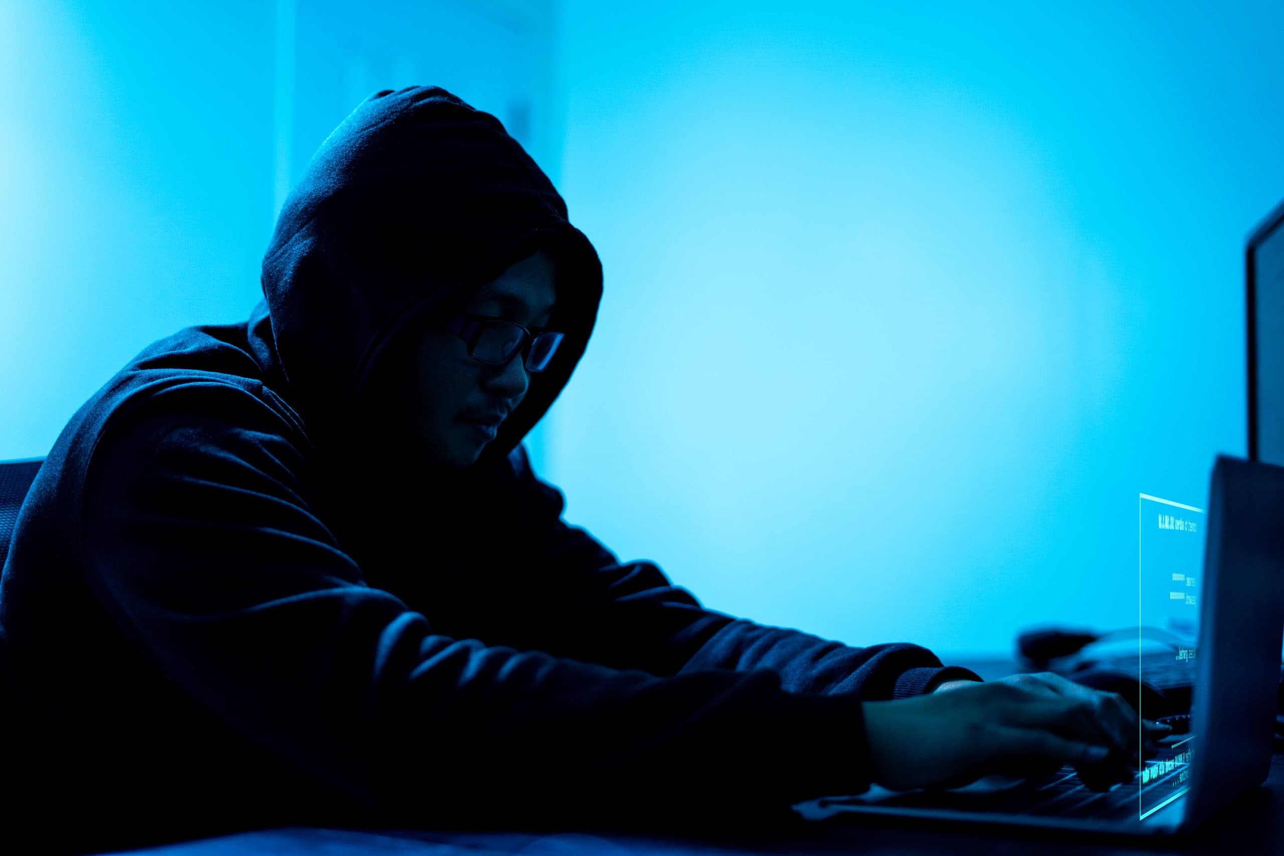 Eine dunkel gekleidete Person sitzt im Schatten vor einem Laptop vor einem blauen Hintergrund