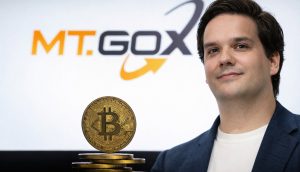 Ein Mann mit dunklem Haar in einem blauen Sakko und weißem T-Shirt steht rechts im Bild vor einem weißen Hintergrund mit dem MT.GOX-Logo. Im Vordergrund sind mehrere gestapelte goldene Bitcoin-Münzen zu sehen, eine davon aufrecht stehend mit gut erkennbarem „B“-Symbol.