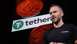 Ein Mann mit kurzem Haar und Bart trägt ein schwarzes Tether-T-Shirt und spricht mit Headset-Mikrofon auf einer Bühne. Hinter ihm ist groß das Tether-Logo zu sehen, davor leuchten rötlich eingefärbte Krypto-Münzen im Hintergrund.