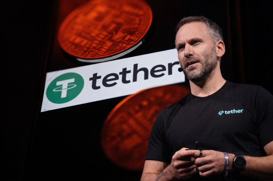 Ein Mann mit kurzem Haar und Bart trägt ein schwarzes Tether-T-Shirt und spricht mit Headset-Mikrofon auf einer Bühne. Hinter ihm ist groß das Tether-Logo zu sehen, davor leuchten rötlich eingefärbte Krypto-Münzen im Hintergrund.