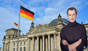 Man sieht das Gebäude des Deutschen Reichstags und davor eine Deutschlandflagge wehen. Rechtsbündig steht ein Mann mit schwarzem Rollkragenpullover.