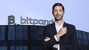 Ein professionell gekleideter Mann im dunklen Anzug steht selbstbewusst mit verschränkten Armen vor einem modernen Bürogebäude aus Glas. Auf dem Dach ist gut sichtbar das „bitpanda“-Logo angebracht, vor einem klaren blauen Himmel.