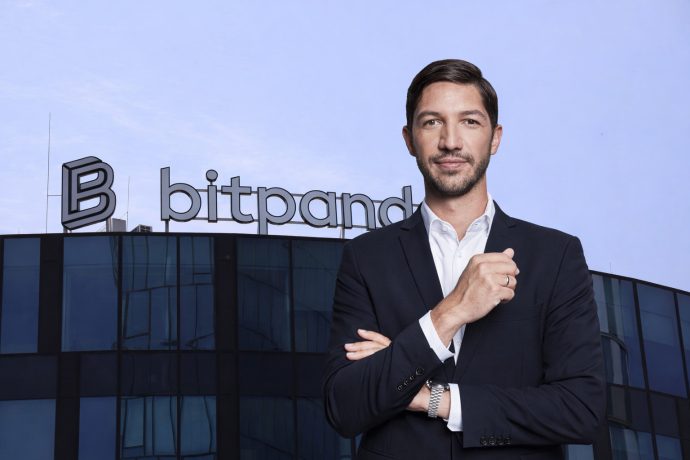 Ein professionell gekleideter Mann im dunklen Anzug steht selbstbewusst mit verschränkten Armen vor einem modernen Bürogebäude aus Glas. Auf dem Dach ist gut sichtbar das „bitpanda“-Logo angebracht, vor einem klaren blauen Himmel.