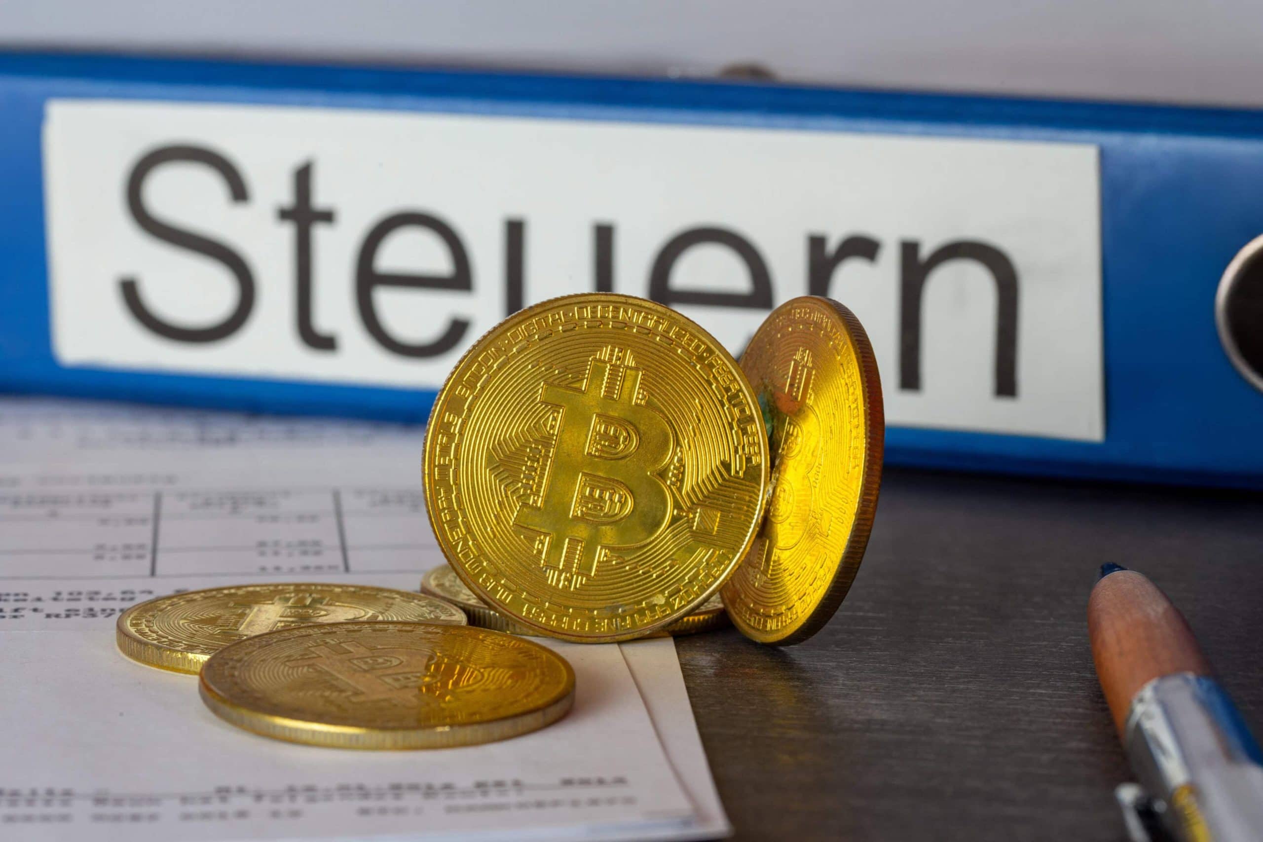 Ein paar vergoldete Bitcoin-Münzen vor einem Aktenordner mit der Aufschrift Steuern.
