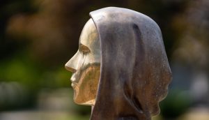 Seitliche Nahaufnahme einer Bronzestatue, die den Kopf und die Schultern einer Person mit Kapuze darstellt, inspiriert von Bitcoin, im Freien mit unscharfem Hintergrund.