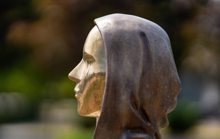 Satoshi Nakamoto ist der Bitcoin-Gründer. Seitliche Nahaufnahme einer Bronzestatue, die den Kopf und die Schultern einer Person mit Kapuze darstellt, inspiriert von Bitcoin, im Freien mit unscharfem Hintergrund.