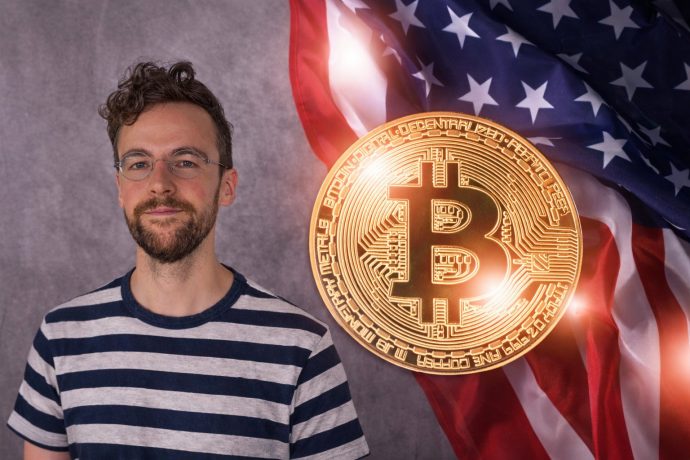 Ein Mann mit lockigen braunen Haaren, Bart und runder Brille steht links im Bild und blickt ruhig in die Kamera. Er trägt ein gestreiftes T-Shirt. Rechts daneben ist eine große, leuchtende Krypto-Münze vor einer wehenden US-Flagge zu sehen.