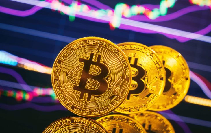 Drei Bitcoin-Münzen stehen aufrecht vor einem bunt gefärbtem Chart als Hintergrund