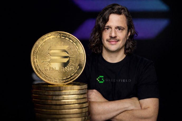 Ein Mann mit schulterlangen dunklen Haaren und Schnurrbart steht mit verschränkten Armen neben einem Stapel goldener Solana-Coins. Er trägt ein schwarzes T-Shirt mit grünem „Greenfield“-Logo. Im Vordergrund steht eine große Solana-Münze auf gestapelten Coins, während der Hintergrund dunkel mit violett-blauen Lichteffekten ist.