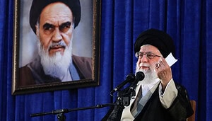 Ayatollah Ali Khamenei bei einer Rede in Teheran vor blauem Vorhang mit Porträt von Ruhollah Khomeini im Hintergrund.