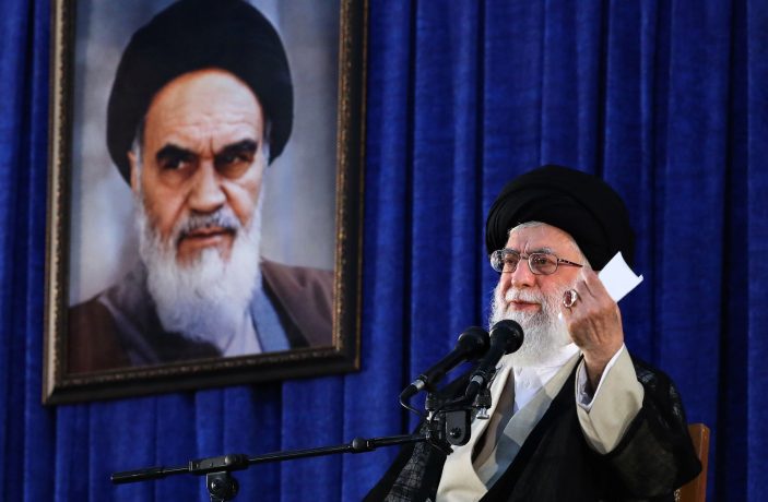 Ayatollah Ali Khamenei bei einer Rede in Teheran vor blauem Vorhang mit Porträt von Ruhollah Khomeini im Hintergrund.