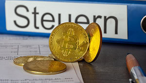 Ein paar vergoldete Bitcoin-Münzen vor einem Aktenordner mit der Aufschrift Steuern.