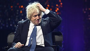 Boris Johnson sitzt auf einer Bühne und trägt einen Anzug