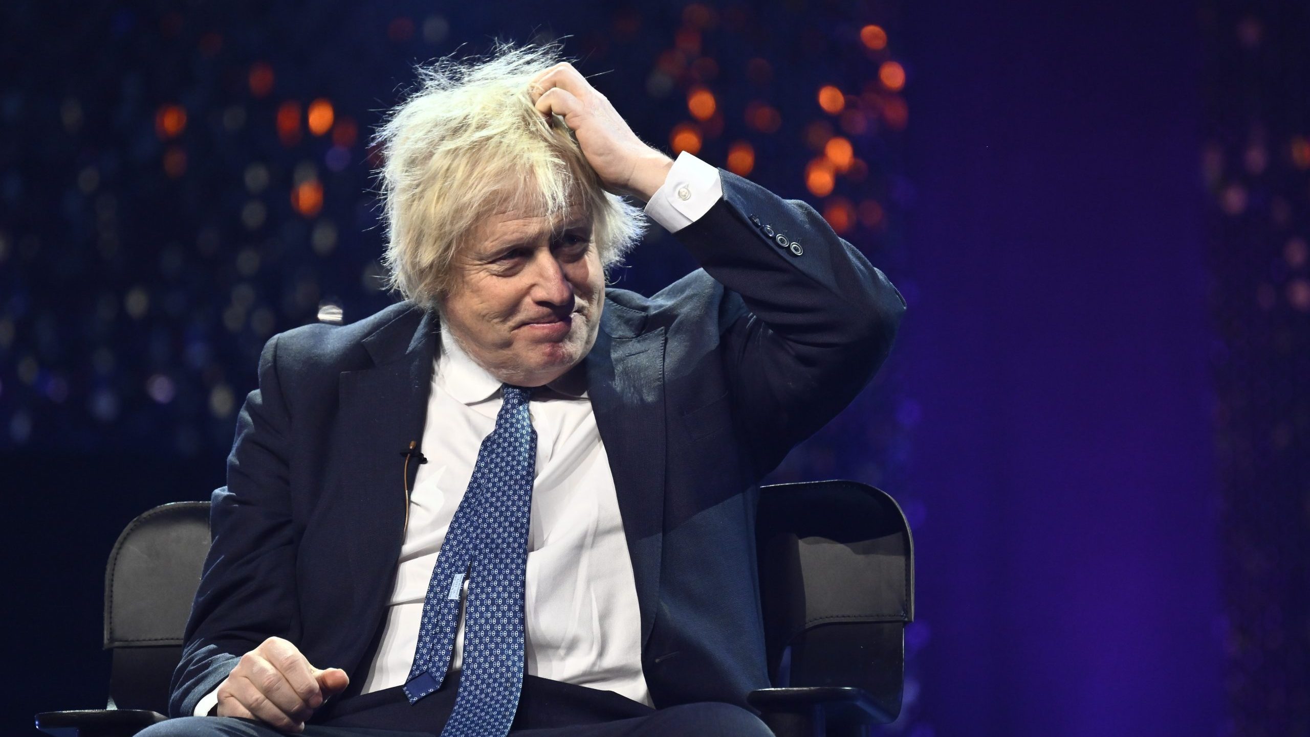 Boris Johnson sitzt auf einer Bühne und trägt einen Anzug