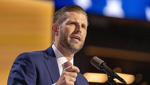 Eric Trump, ein Mann in einem blauen Anzug und einer rot gestreiften Krawatte, der Trump ähnelt, spricht auf einer Bühne in ein Mikrofon, vor einem verschwommenen Hintergrund mit blauen und gelben Lichtern.