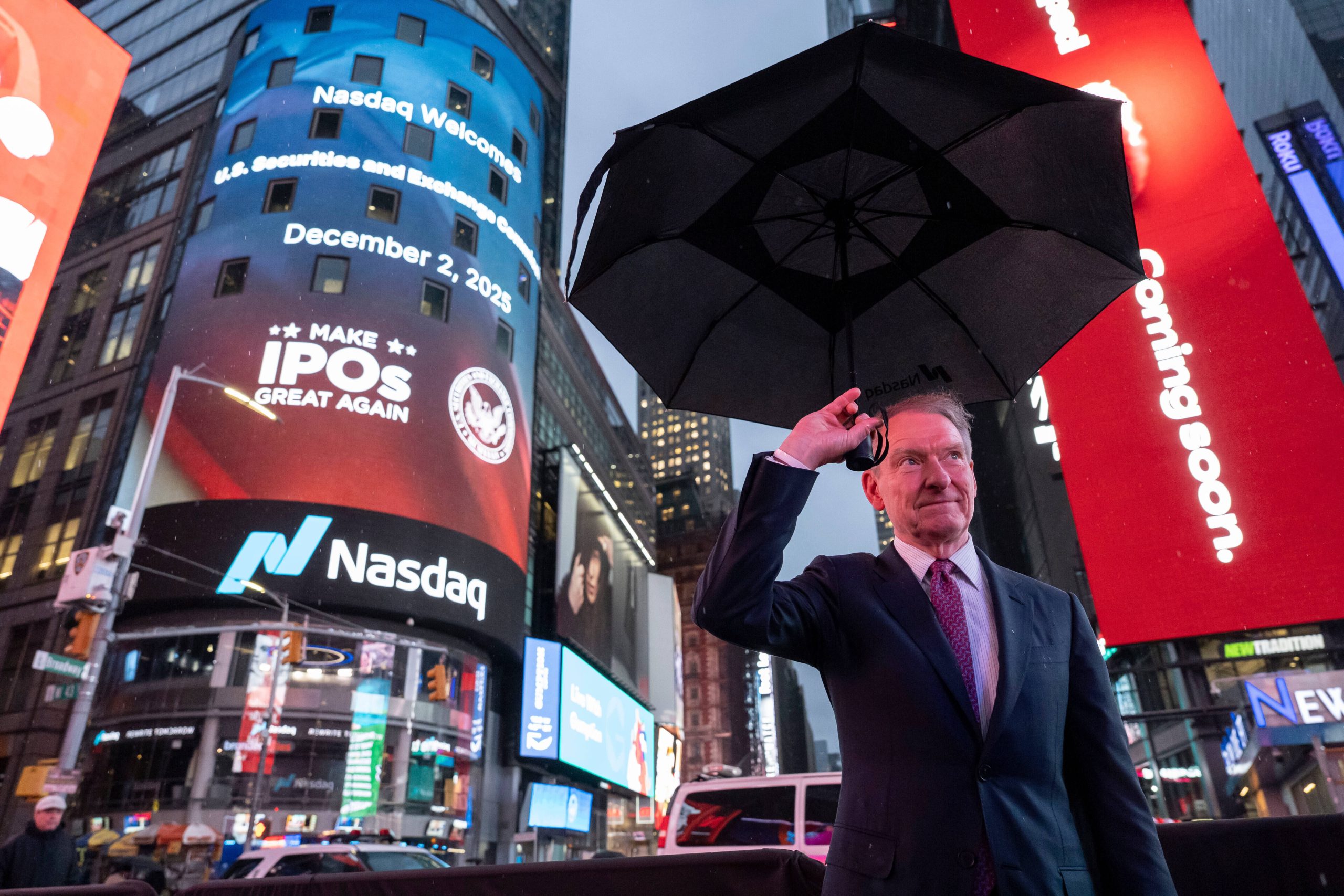 Das Bild zeigt eine Straßenszene in New York am Times Square bei Regen. Im Vordergrund steht ein Mann im Anzug, der einen aufgespannten schwarzen Regenschirm über sich hält.