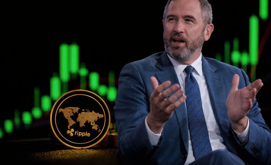 Ein Mann mittleren Alters mit kurzen grauen Haaren und grauem Bart trägt einen blauen Anzug und gestikuliert mit den Händen, als würde er etwas erklären. Vor ihm liegen goldene Ripple-Coins mit Logo, während im Hintergrund ein unscharfer grüner Kryptowährungs-Candlestick-Chart auf schwarzem Hintergrund zu sehen ist.